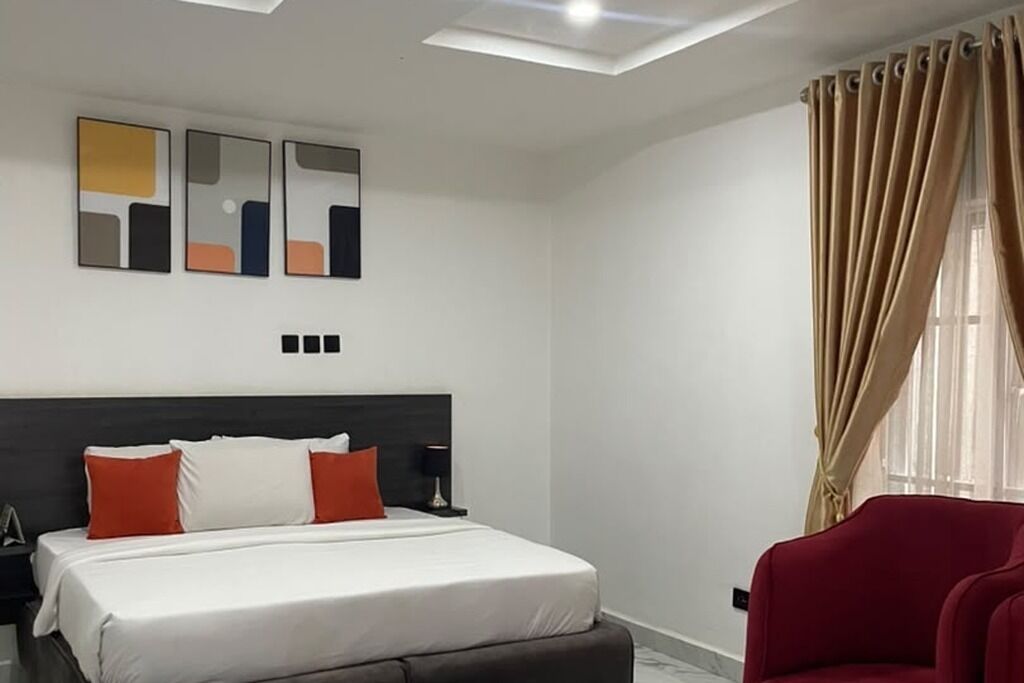 Une chambre d’hôtel moderne avec un lit, un fauteuil rouge, des œuvres d’art abstraites accrochées au mur et une fenêtre avec des rideaux.