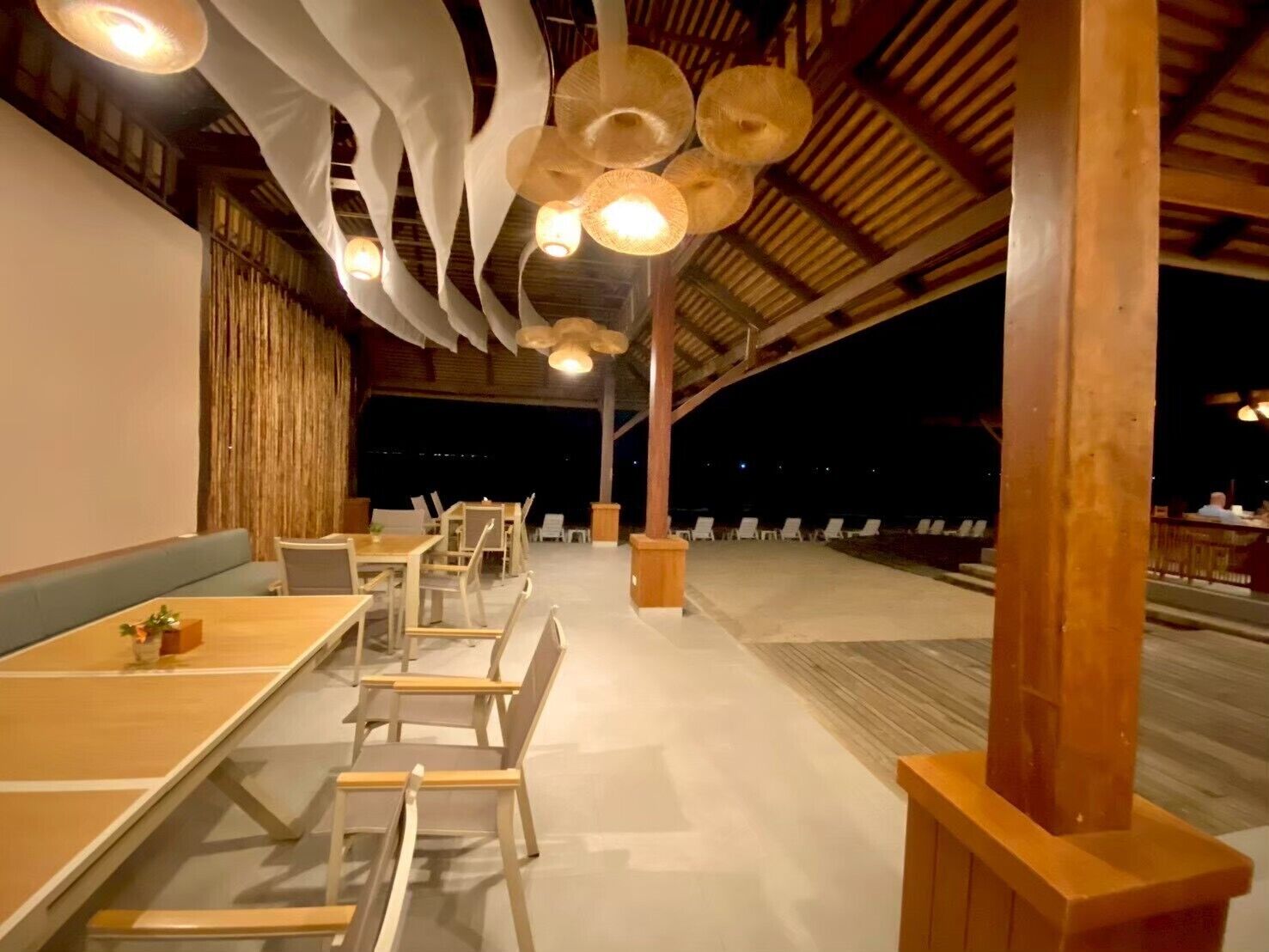 A restaurant with long wooden tables and chairs, a high ceiling with hanging light fixtures, and a view of the beach at night.