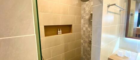 Un baño con ducha, tocador con espejo y estante para artículos de aseo.