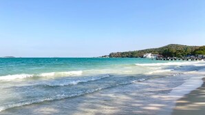 Una playa con agua cristalina, una orilla arenosa y una colina con edificios al fondo.