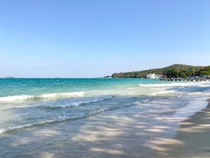 Una playa con agua cristalina, una orilla arenosa y una colina con edificios al fondo.