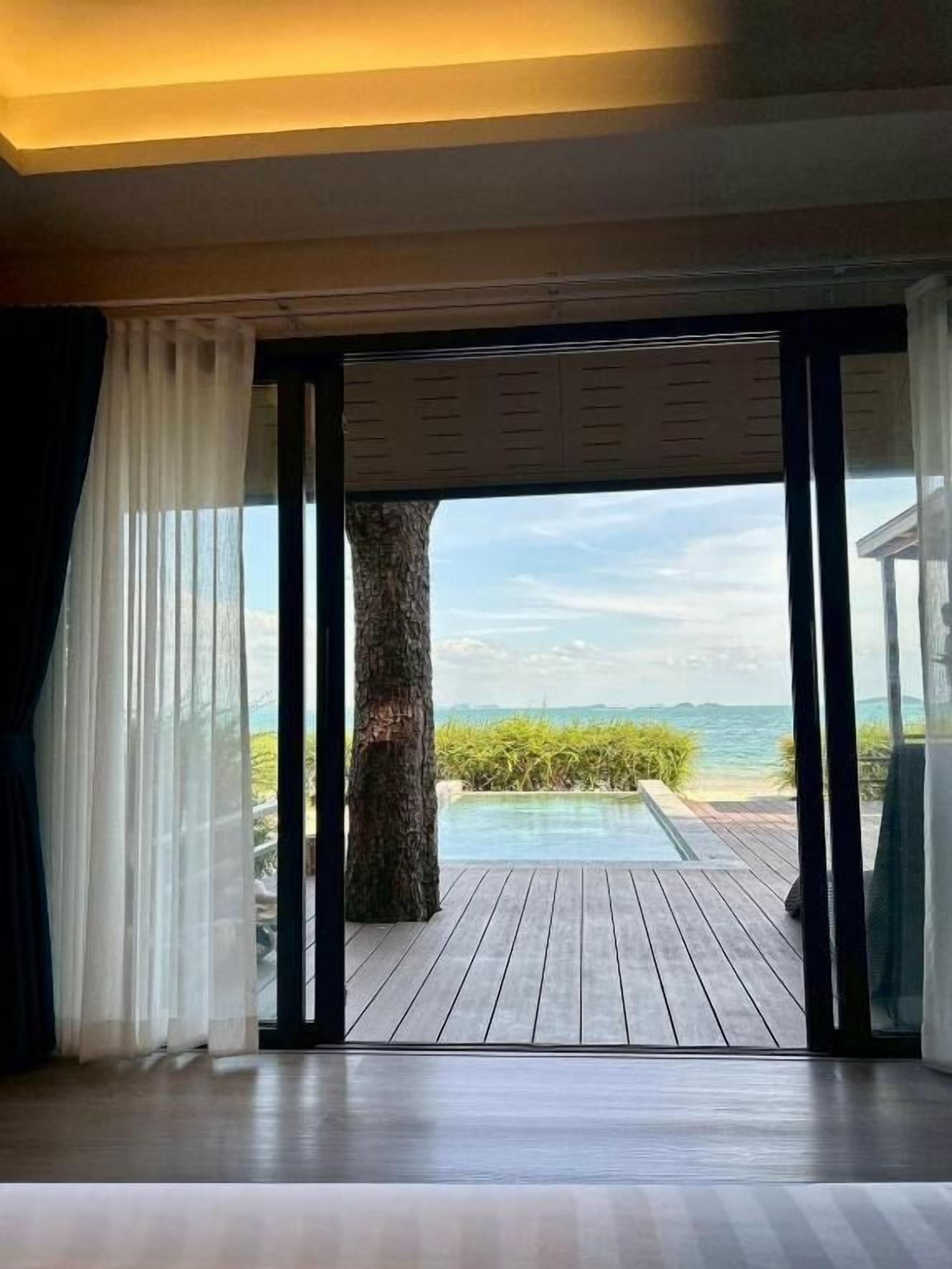 Una habitación con un amplio ventanal corredera que da a una terraza de madera con piscina y vistas al océano.