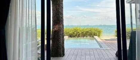 Una habitación con un amplio ventanal corredera que da a una terraza de madera con piscina y vistas al océano.