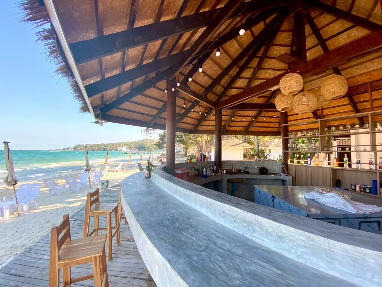 Un bar junto a la playa con sillas de madera y una barra, con vista al océano.