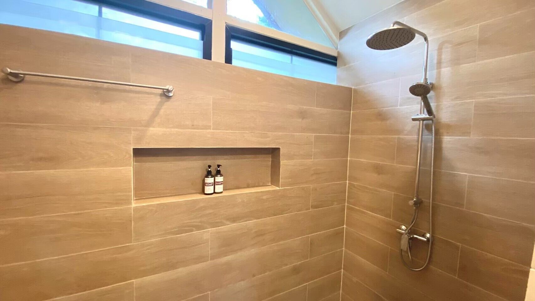 A shower area with a handheld showerhead and a wall-mounted faucet. There is a shelf with two bottles of soap and shampoo.
