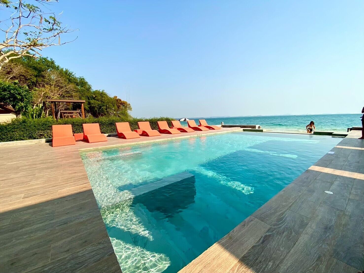 A swimming pool with clear blue water, surrounded by wooden deck and red lounge chairs.