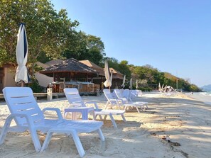 Un resort frente al mar con sillones de playa y sombrillas, un área de bar y una construcción con techo de paja.