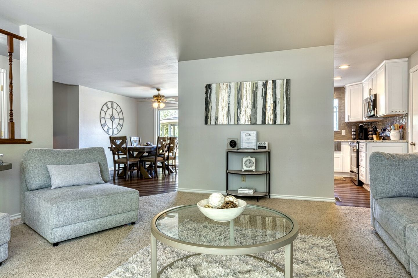 A modern living room with a glass coffee table, grey sofas, a dining area with a clock, and a kitchen in the background.