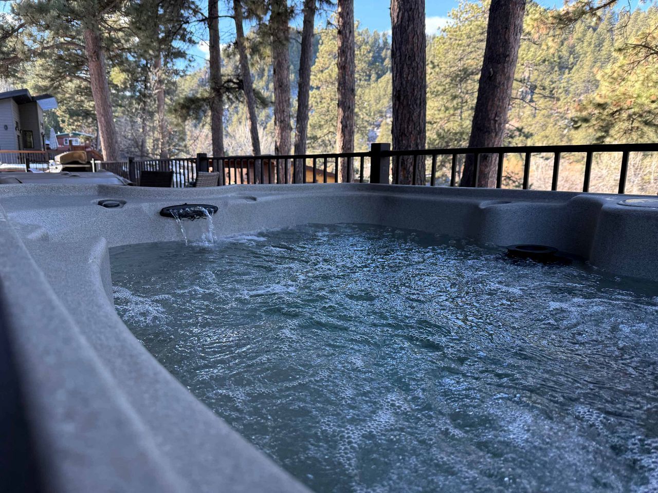 A hot tub with water flowing, surrounded by a wooden deck and a metal railing, with trees in the background.