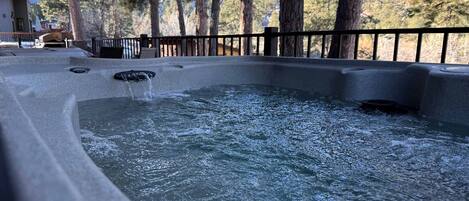 A hot tub with water flowing, surrounded by a wooden deck and a metal railing, with trees in the background.
