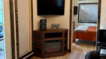 A room with a flat-screen TV mounted on the wall, a small wooden entertainment unit with a fireplace, a bed with an orange bedspread, and a black leather chair.