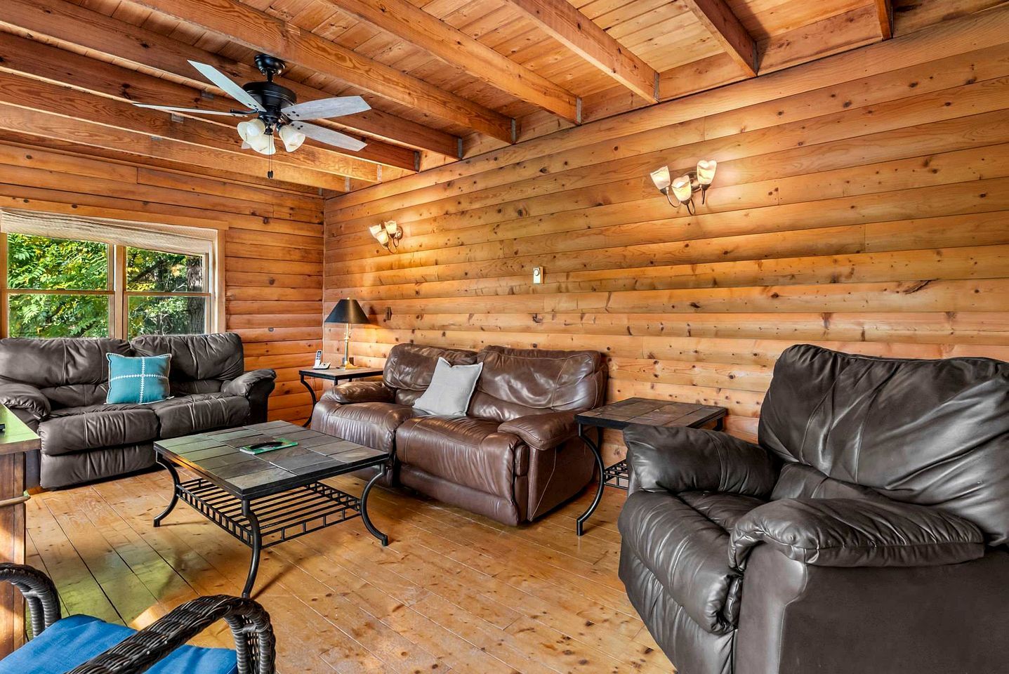 A log cabin interior with leather sofas, a coffee table, a ceiling fan, and wall-mounted lights.