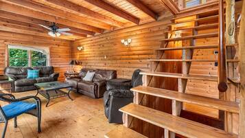 A log cabin interior with a fireplace, wooden shelves, leather sofas, and a ceiling fan.
