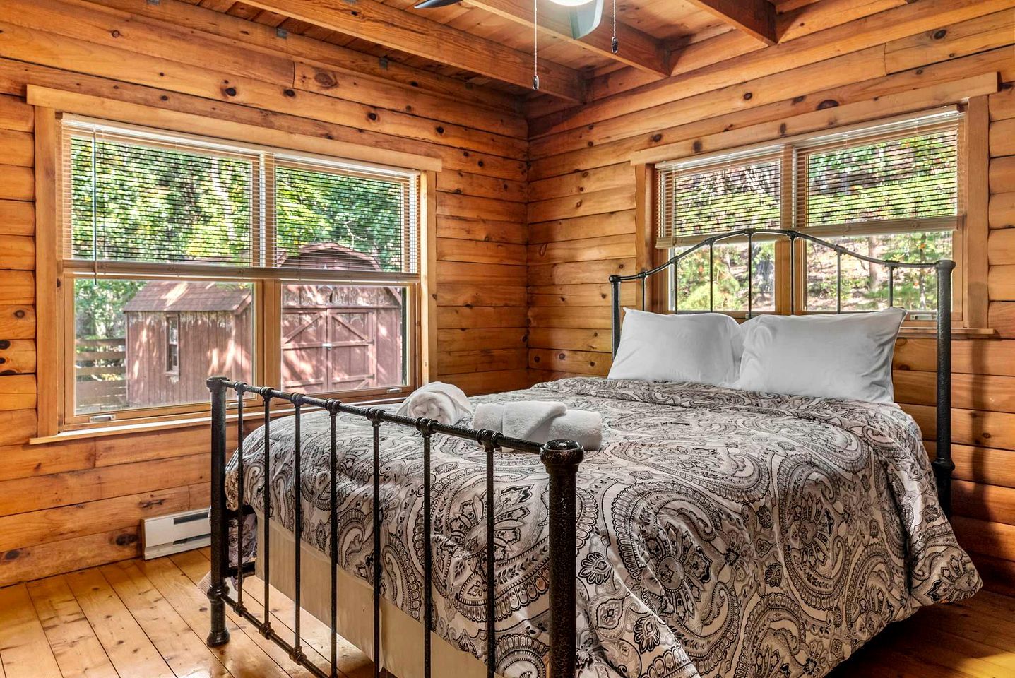 Un dormitorio de cabaña de madera con una cama, dos ventanas y vista a una construcción de madera al exterior.