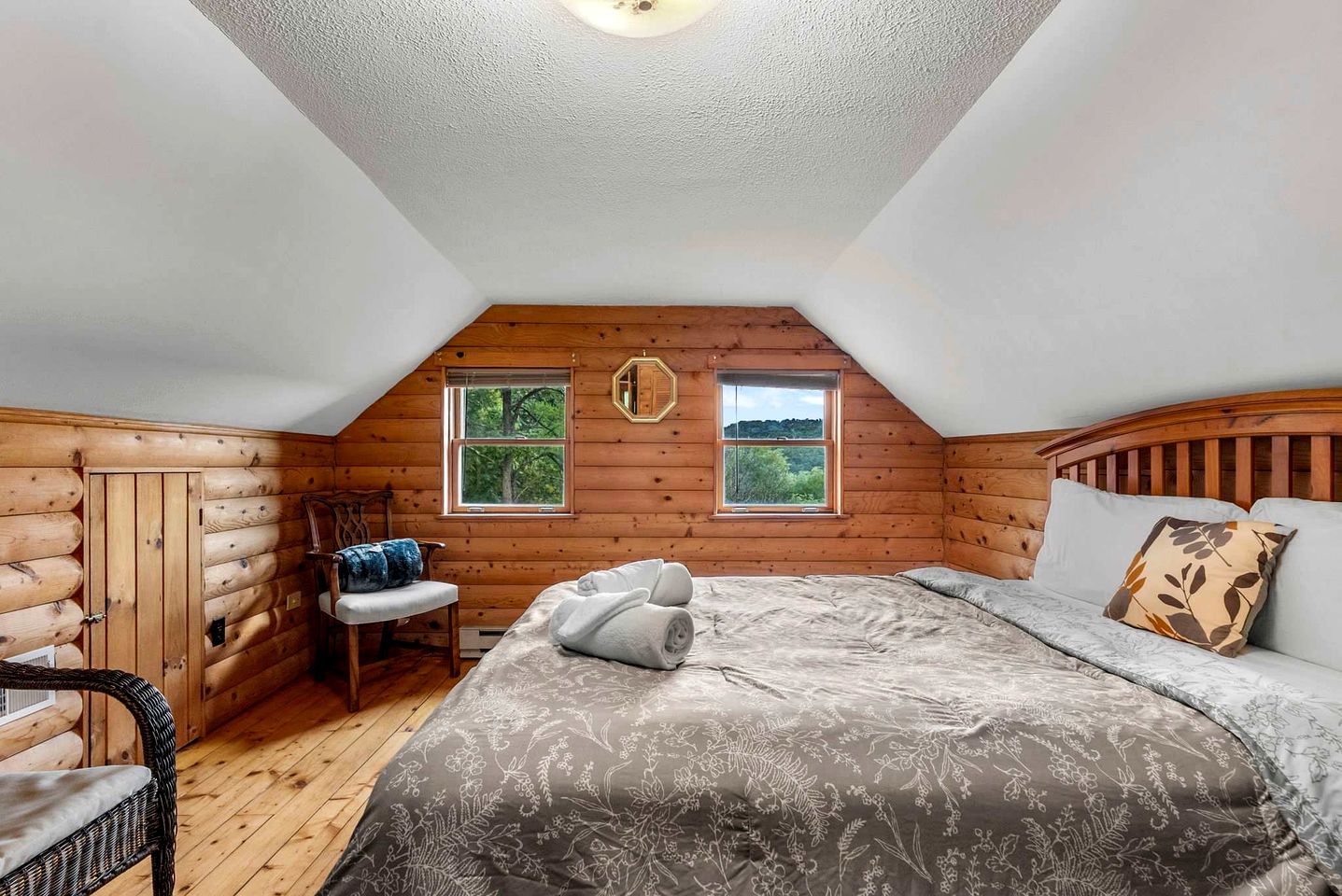 Een slaapkamer in een blokhut met een bed, twee ramen, een stoel en een houten hoofdbord.