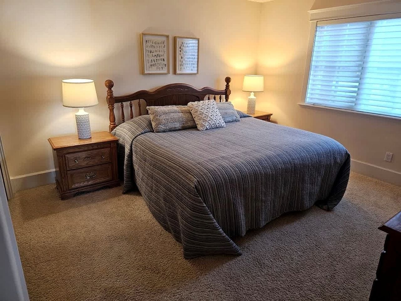 A bedroom with a bed, two bedside tables, a nightstand, a window with blinds, and two wall-mounted pictures.