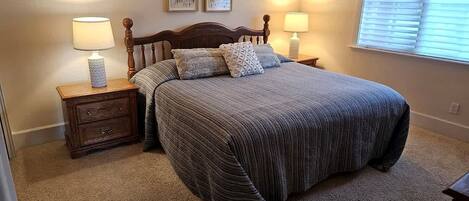 A bedroom with a bed, two bedside tables, a nightstand, a window with blinds, and two wall-mounted pictures.