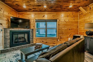 A log cabin interior with a stone fireplace, leather sofa, and a television mounted on the wall. - Stylish Pigeon Forge Cabin with Hot Tub & Resort Amenities near Gatlinburg Golf Course, TN (Pigeon Forge)