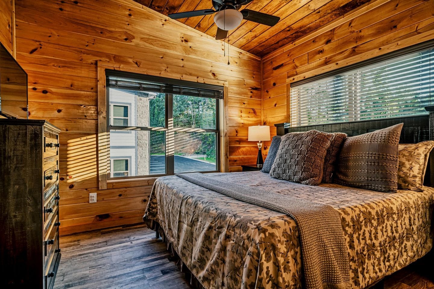 A wooden cabin bedroom with a bed, dresser, and a window overlooking a building.