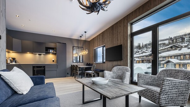 A modern living room with a sofa, a patterned armchair, a coffee table, and a view of a snowy village through large windows.