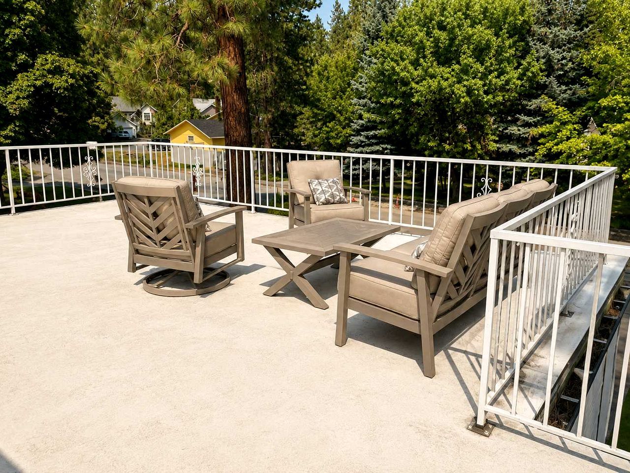 An outdoor patio area with a white metal fence, beige outdoor furniture, and a view of a residential area with trees and houses.
