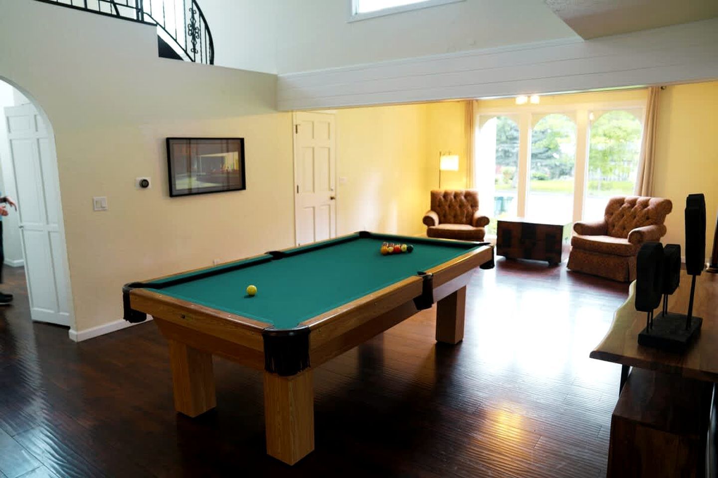A pool table in a room with a staircase, two armchairs, and a door.