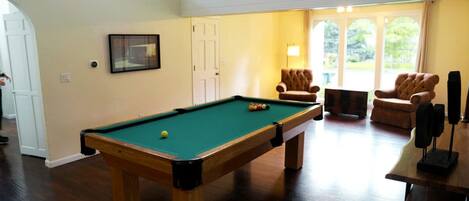 A pool table in a room with a staircase, two armchairs, and a door.