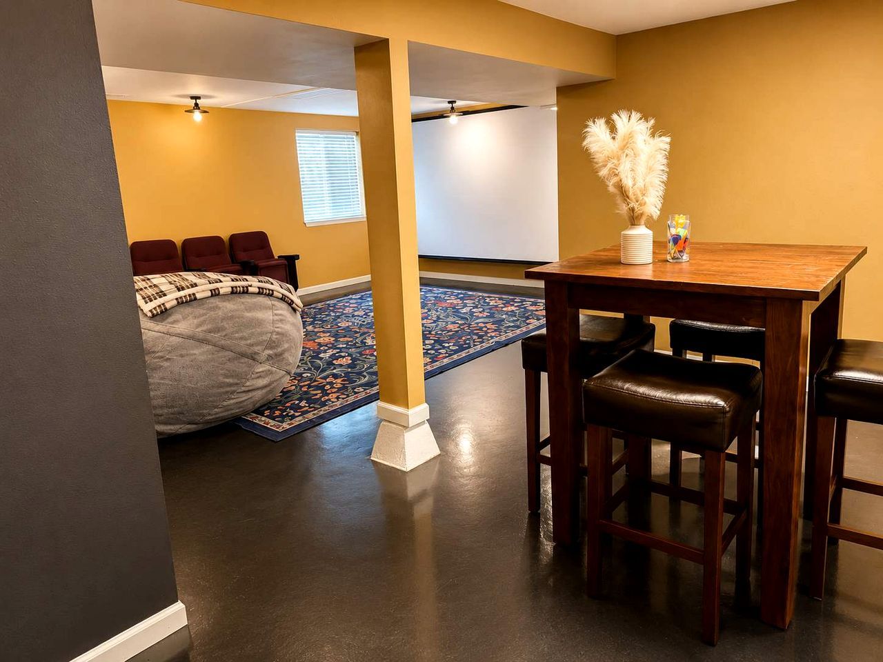 A room with a dark floor, a wooden table with chairs, a bed with a patterned blanket, and a decorative vase with pampas grass.