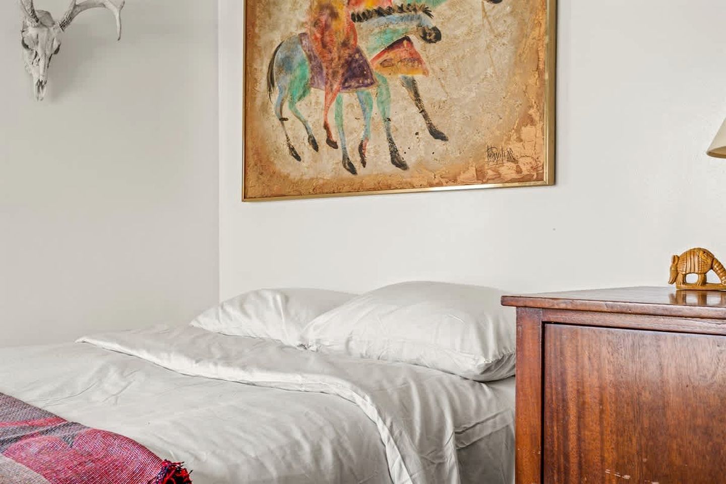 A bedroom with a bed, a wooden nightstand, a skull decoration, and a painting of a horse.