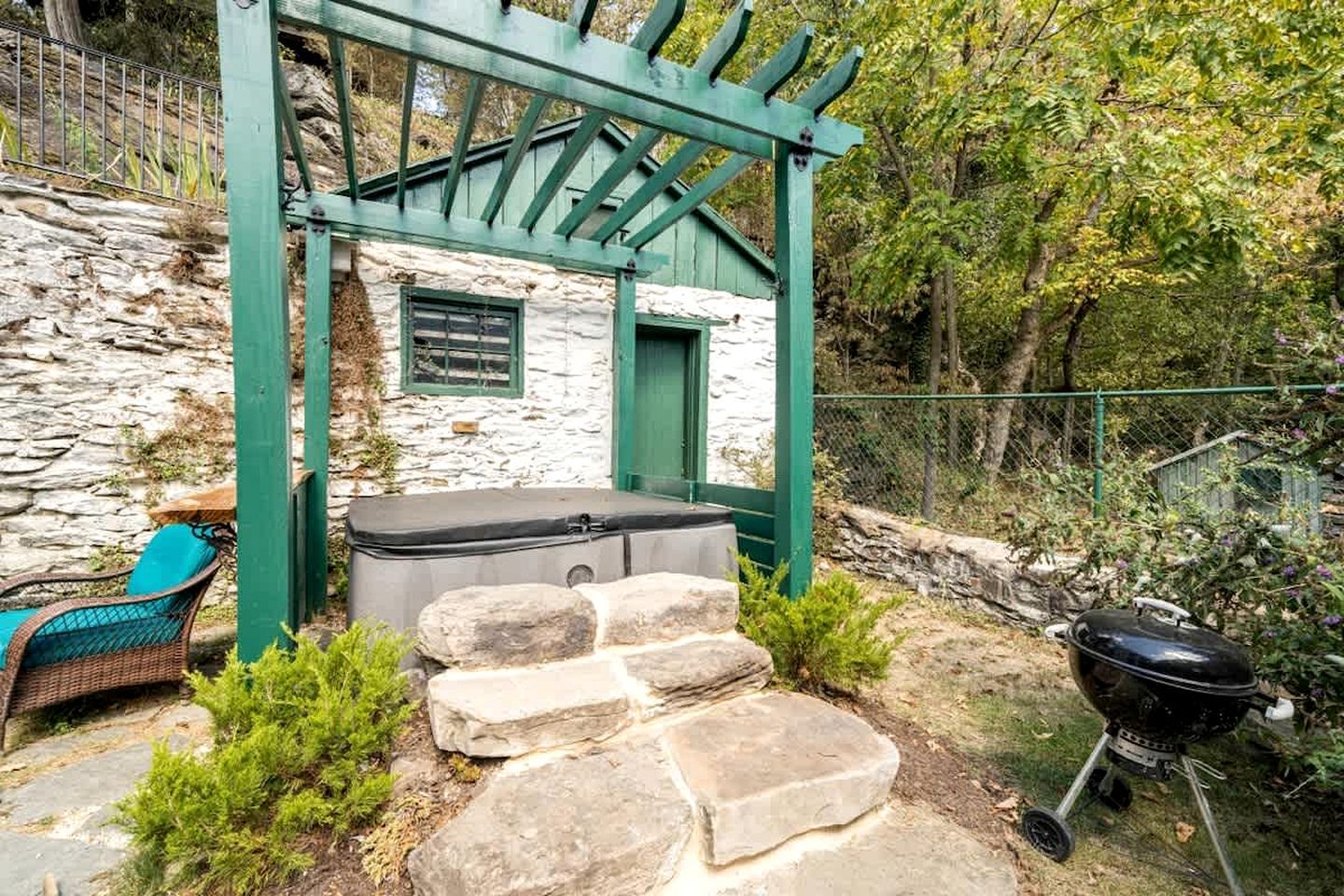 Ein Steinhaus mit einer grünen Pergola, einem Whirlpool, einem Grill und einem Korbstuhl.