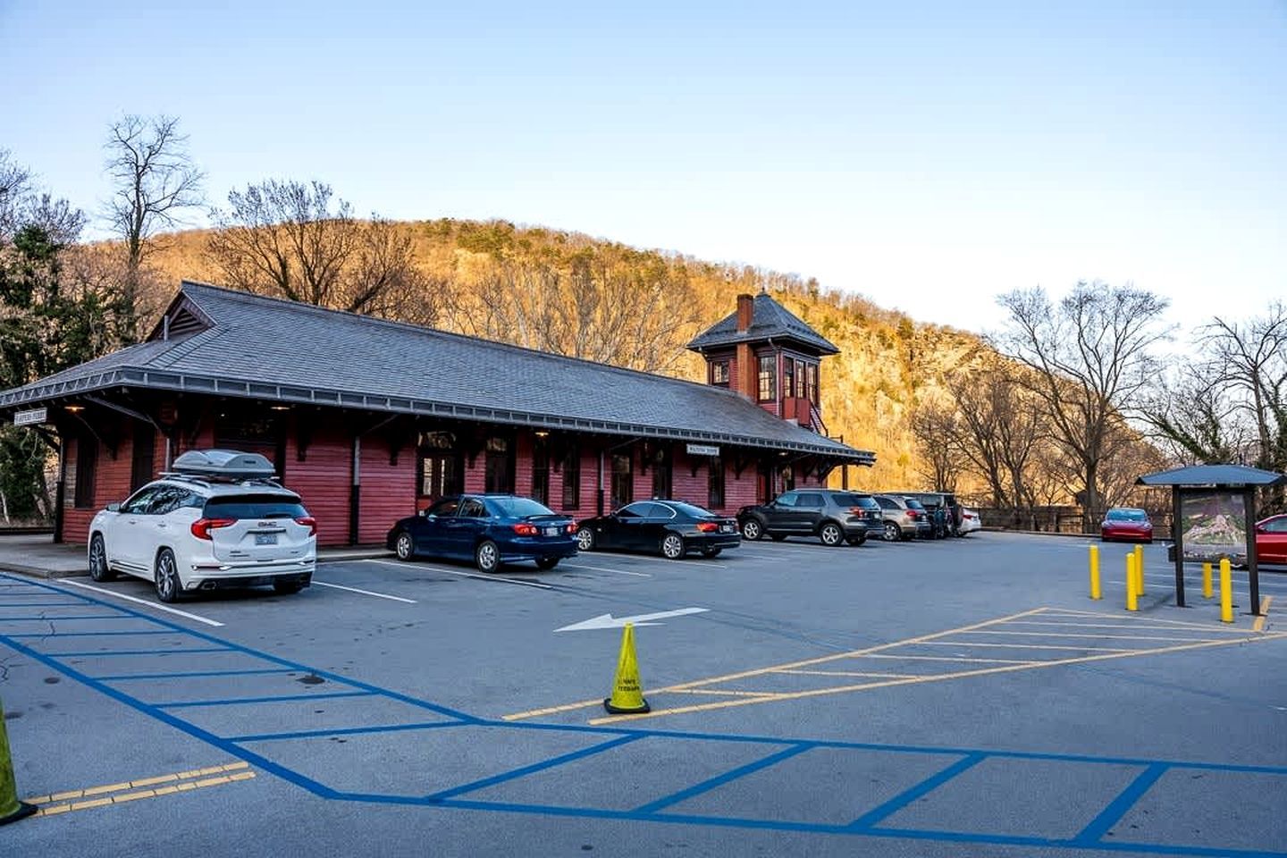 Un edificio de fachada roja y una torre, estacionamiento con espacios delimitados, y un paisaje montañoso de fondo.