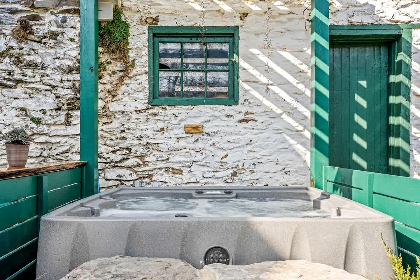 A stone building with a green door and window, a hot tub, and a stone wall.