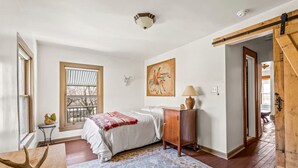 A bedroom with a bed, a window, a painting, a lamp, and a wooden door.