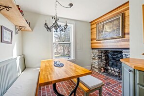 Uma cozinha com mesa de madeira, banco, lareira de pedra e um candelabro pendurado.