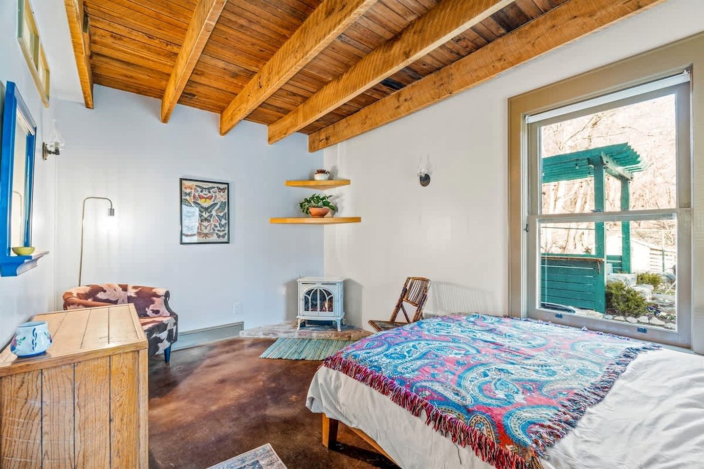 Een slaapkamer met een houten plafond, een bed met een kleurrijk dekbed, een stoel, een klein tafeltje, een wandplankje met een plant, en een raam met uitzicht.