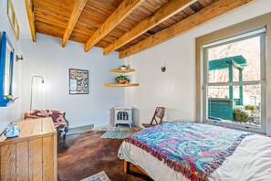 A bedroom with a wooden ceiling, a bed with a colorful blanket, a chair, a small table, a wall-mounted shelf with a plant, and a window with a view of the outside.