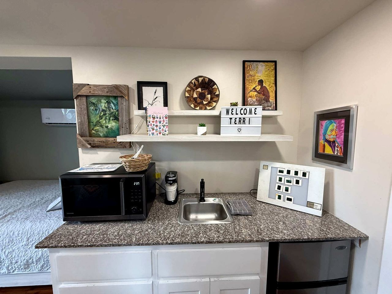 A compact kitchenette with a microwave, coffee maker, and a sink. There are decorative items on the shelves and a framed welcome sign.