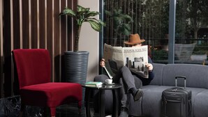 A person sitting on a grey sofa reading a newspaper, with a red chair and a small table with a book and a cup beside them. - Aspera Hotel Taksim (Istanbul)