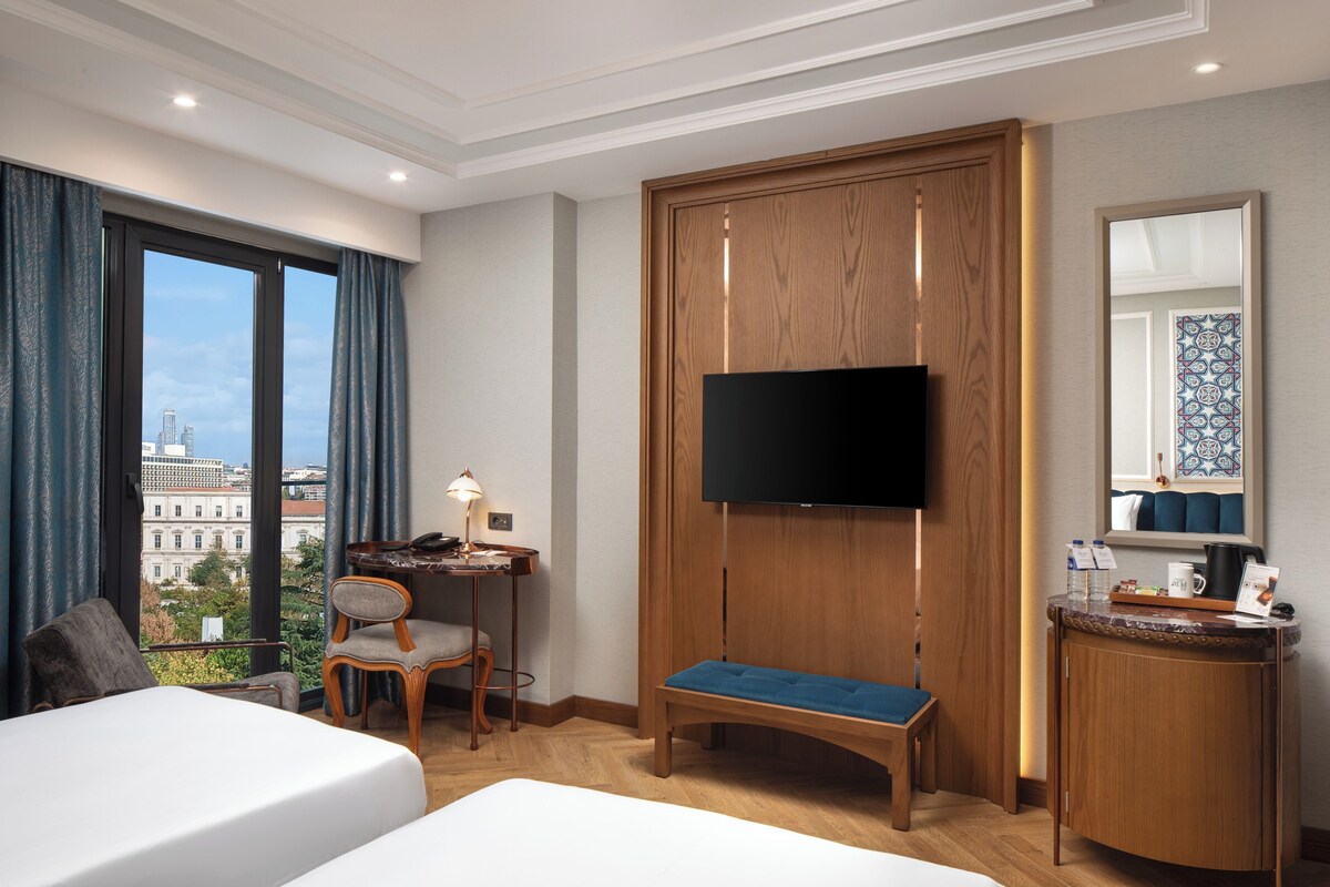Habitació d'hotel amb un llit, un escriptori amb cadira, una televisió muntada sobre un panell de fusta i vistes a la ciutat a través de grans finestres.