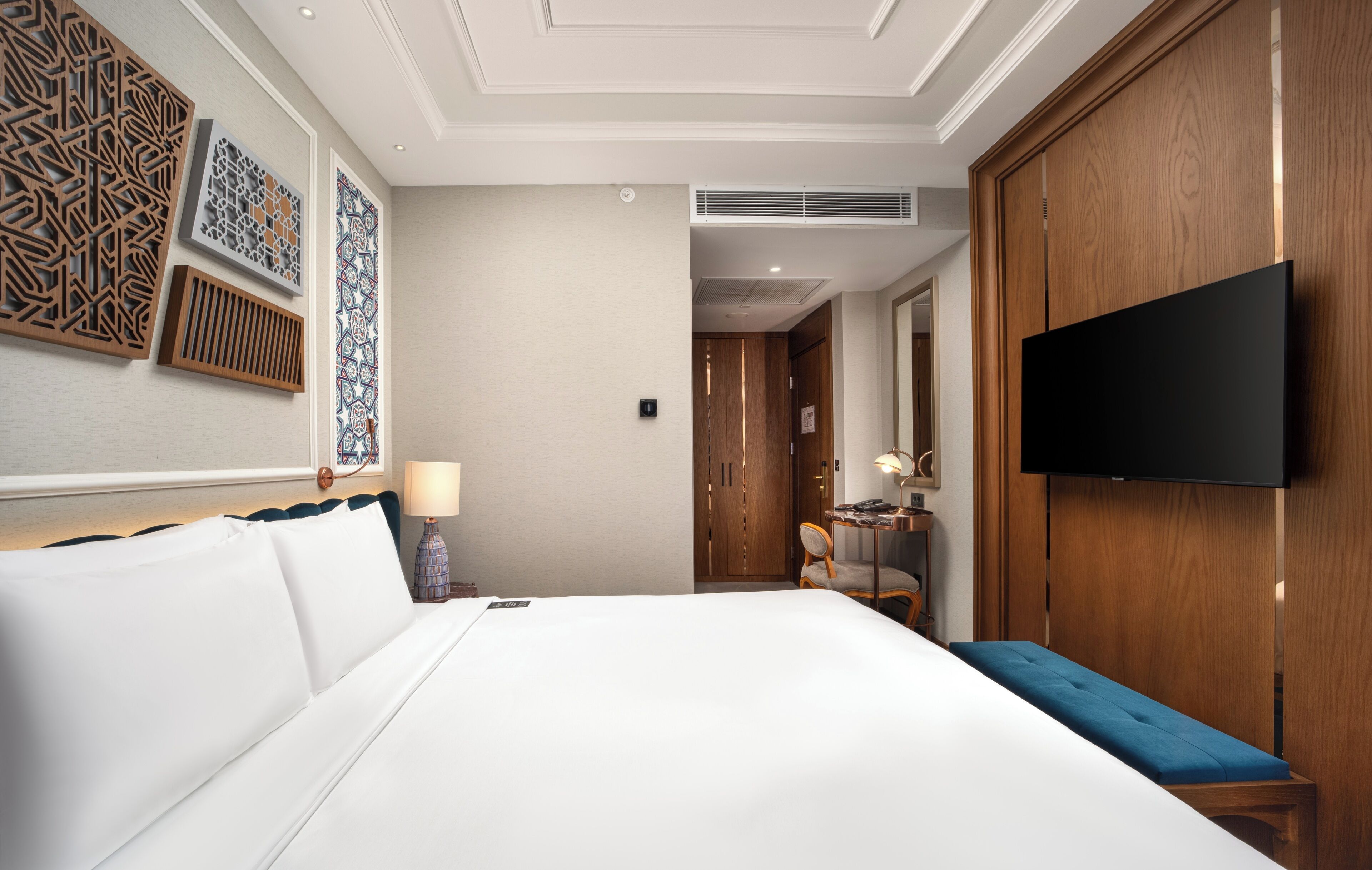A hotel room with a large bed, a flat-screen TV mounted on the wall, a wooden wardrobe, and decorative wall panels.