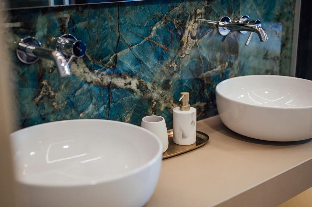 Une salle de bain avec deux lavabos blancs, un pare-baignoire en marbre et un plateau doré.
