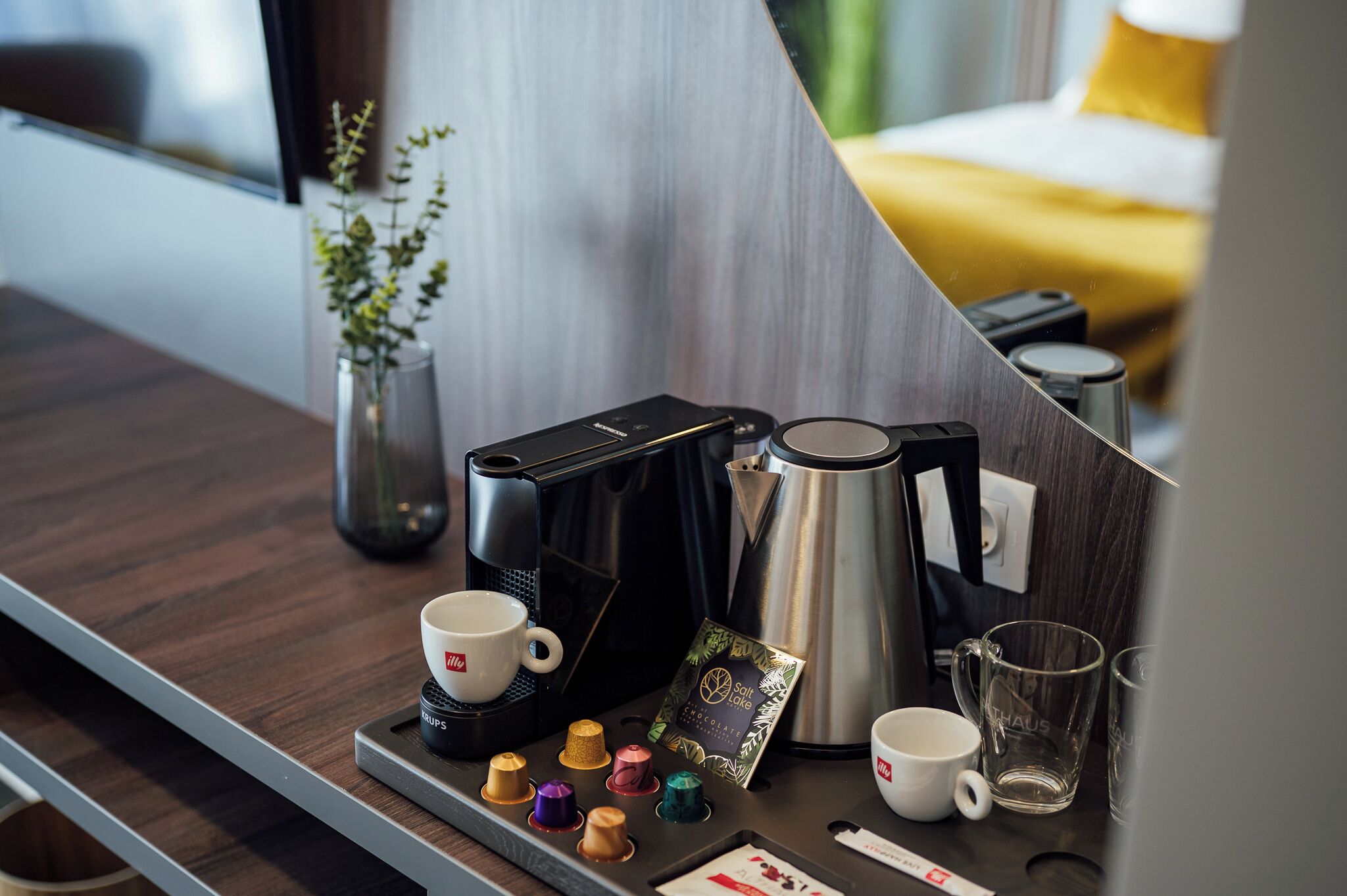 Una estación de café con cafetera, un hervidor, una bandeja con cápsulas de café, tazas y un vaso.