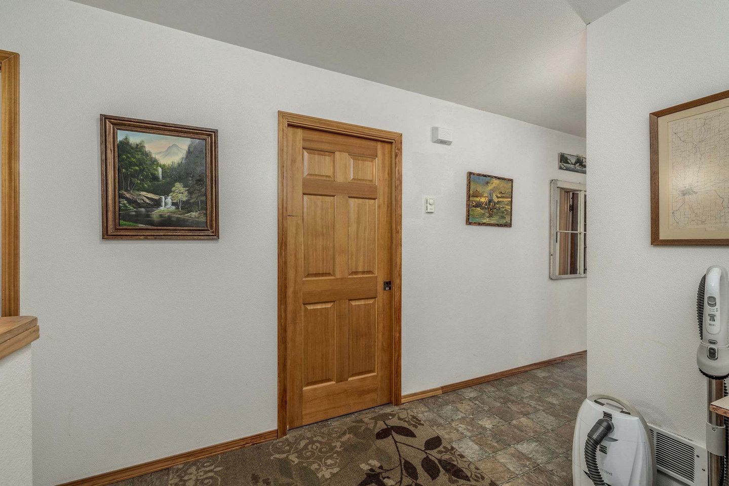 Un pasillo con puerta de madera, cuadros enmarcados y una aspiradora.