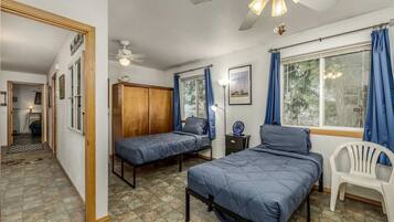 A bedroom with two single beds, a wooden wardrobe, a ceiling fan, a desk with a lamp, and a window with curtains.