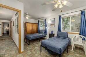 A bedroom with two single beds, a wooden wardrobe, a ceiling fan, a desk with a lamp, and a window with curtains.