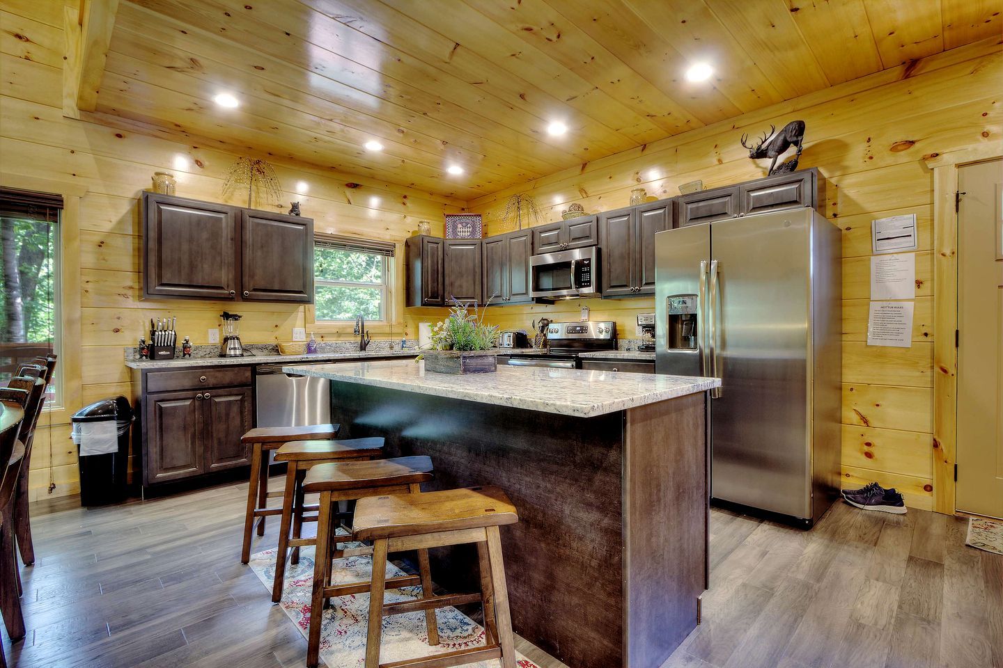A wooden cabin kitchen with dark cabinets, stainless steel appliances, a central island with stools, and a window overlooking greenery.