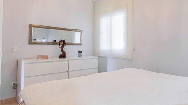 Een slaapkamer met een bed, een dressoir, een spiegel en een raam met jaloezieën.