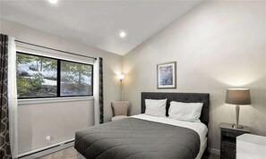 A bedroom with a bed, a nightstand, a lamp, a window with curtains, and a wall-mounted picture.