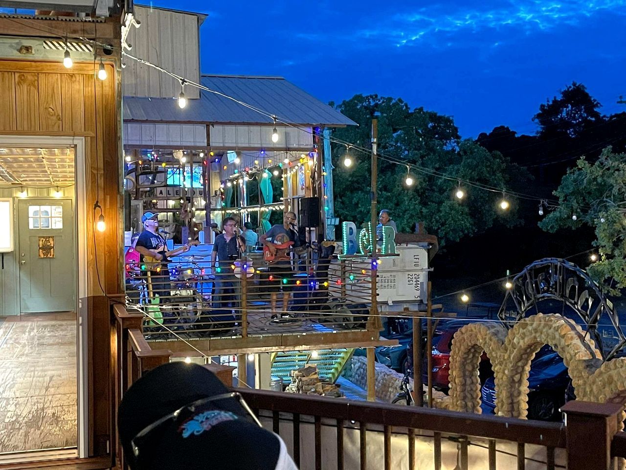 A bar with a stage and seating area, surrounded by string lights and trees.