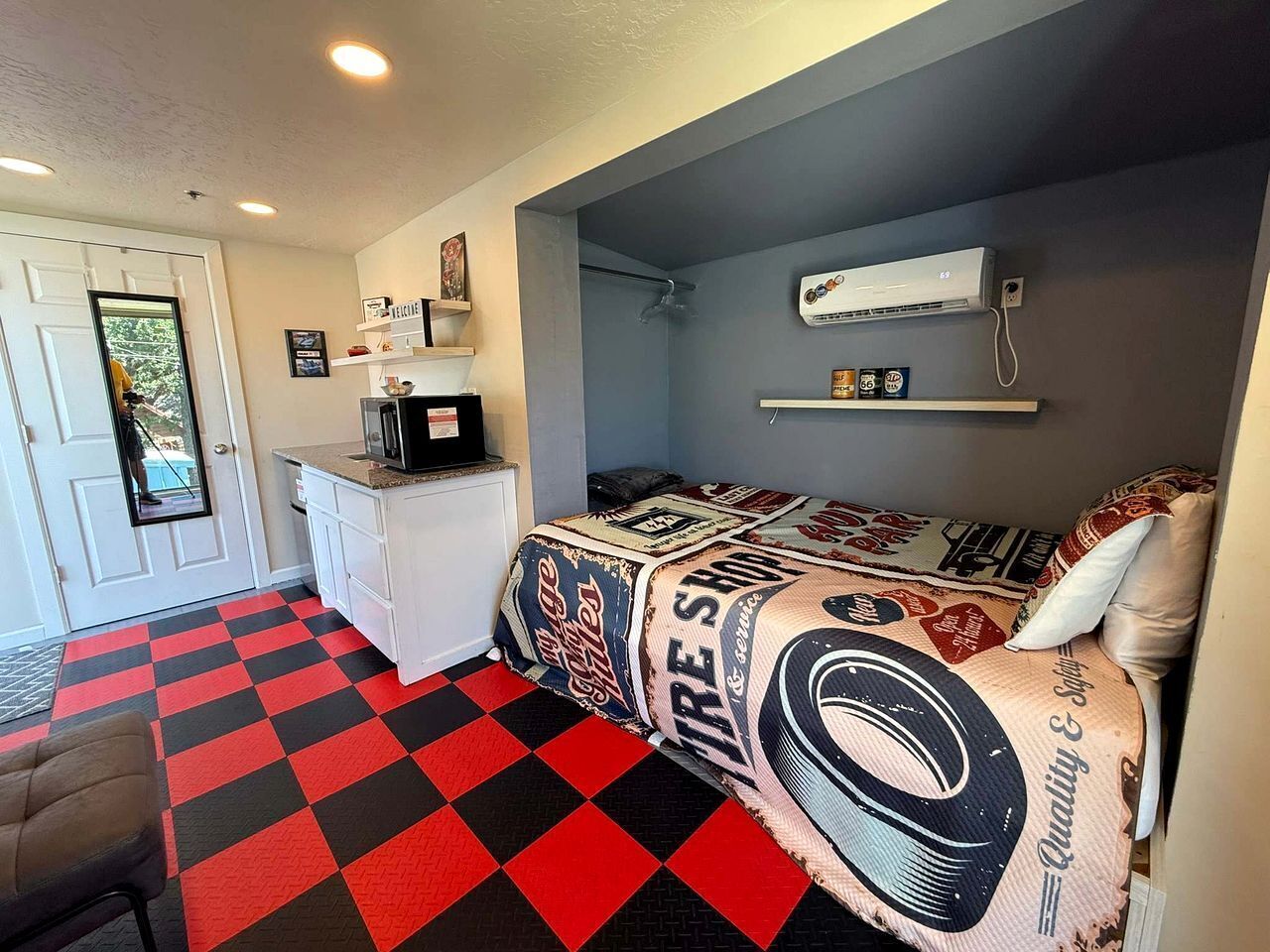 Una habitación compacta con piso a cuadros, una cama con cubrecama con motivos de fuego, un tocador blanco, un microondas y una ventana pequeña.